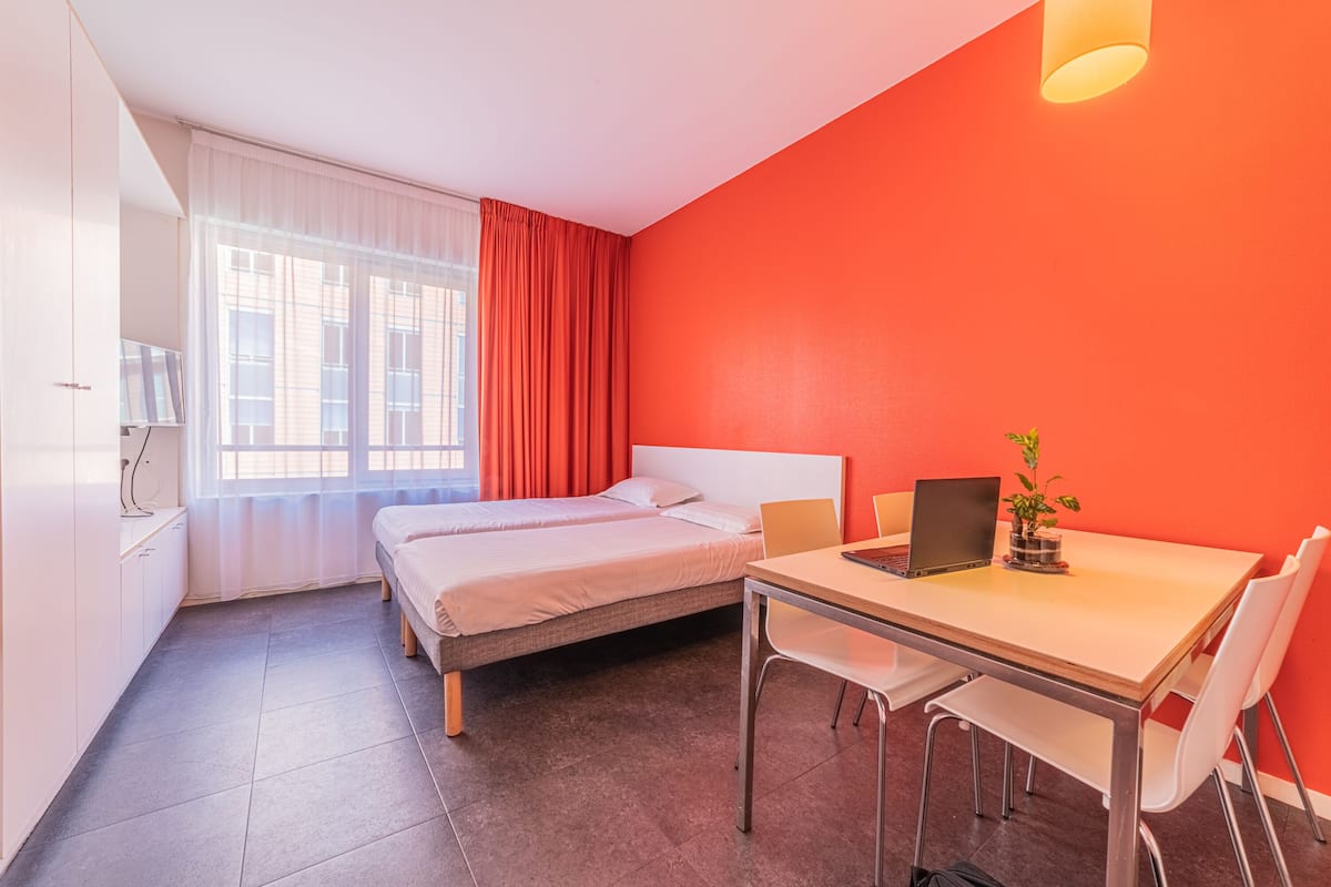 Une chambre avec deux lits, un bureau avec un ordinateur portable, une chaise et une fenêtre avec des rideaux.