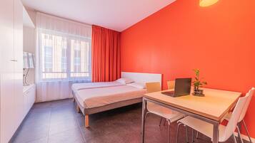 A room with two beds, a desk with a laptop, a chair, and a window with curtains.