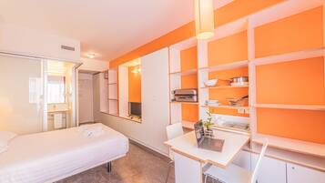A hotel room with a bed, a desk with a laptop, and orange shelving units.