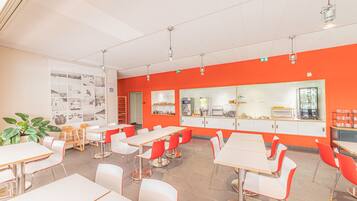 Un espace repas moderne avec des tables blanches et des chaises rouges, un comptoir de cuisine et un mur orné d’un tableau d’affichage.