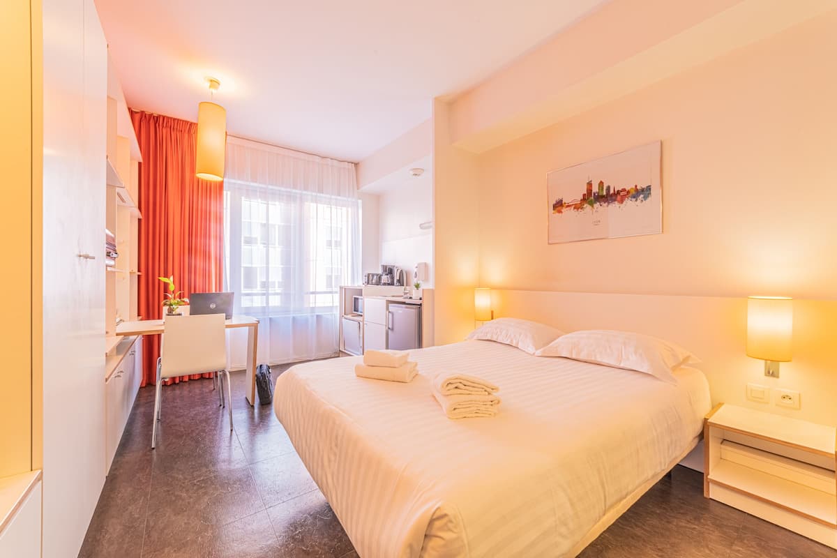 Une chambre d’hôtel équipée d’un lit, d’un bureau, d’une kitchenette et ornée d’une œuvre d’art représentant un paysage urbain au mur.