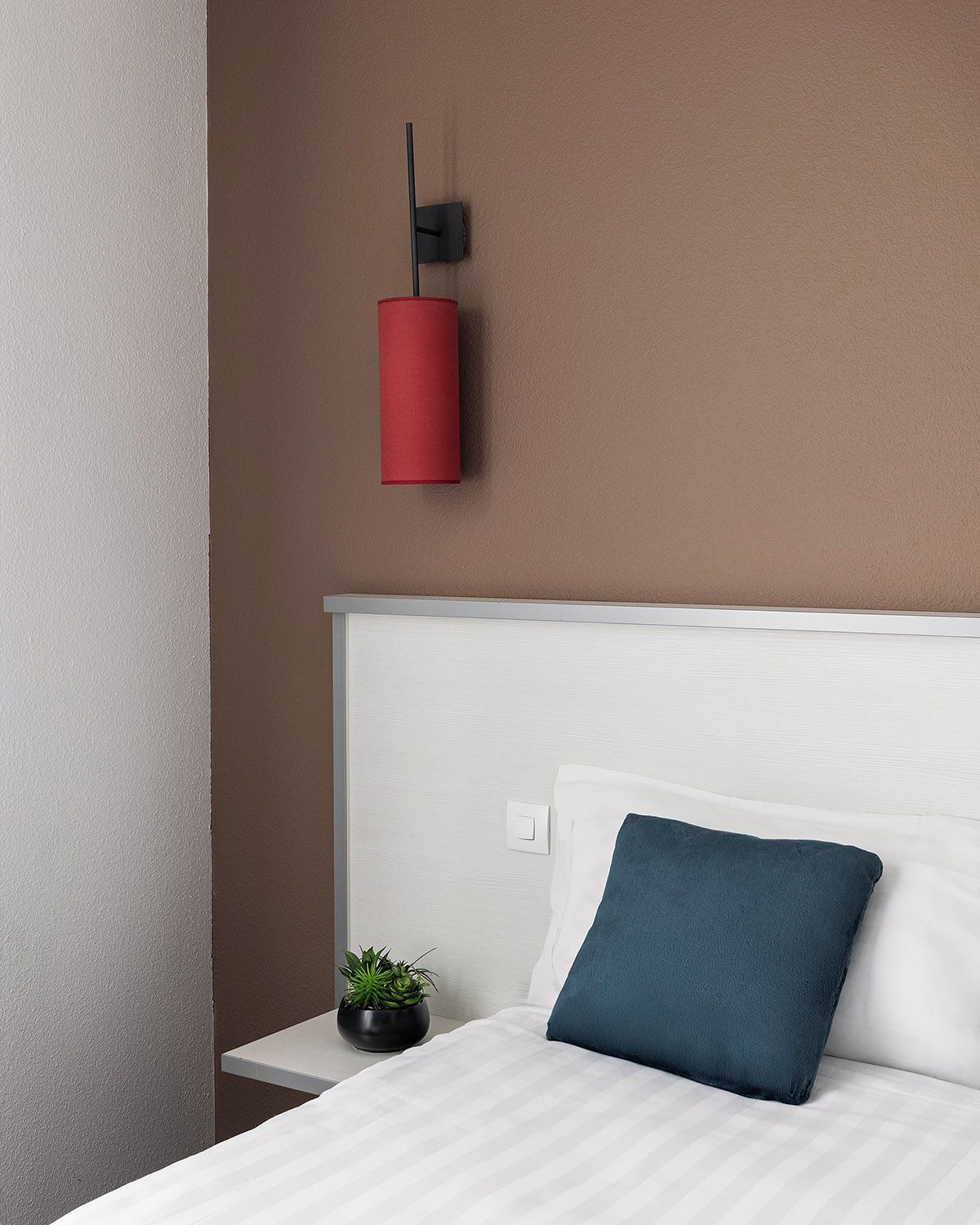 A bedroom with a white bed, a blue pillow, a red wall-mounted light, and a small plant on a nightstand.