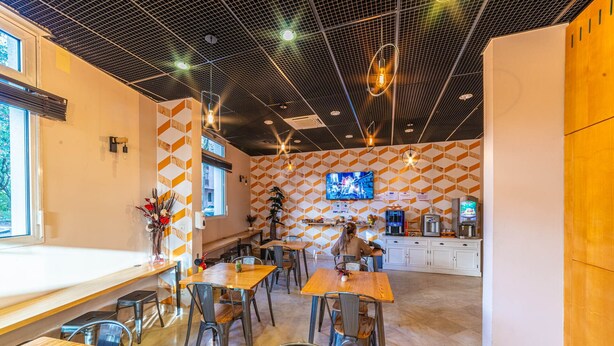 A modern cafe with multiple wooden tables and chairs, a TV mounted on the wall, and a coffee machine. - Montempô Lyon Part Dieu (Lyon)