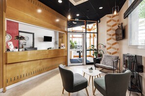 A reception area with a wooden reception desk, two grey chairs, a small table, and a TV mounted on the wall. - Montempô Lyon Part Dieu (Lyon)