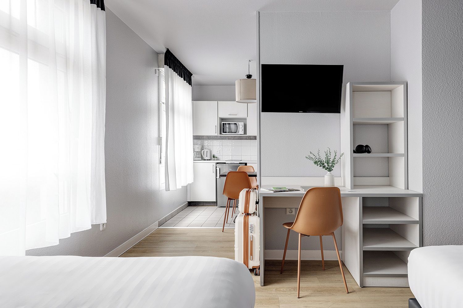 A modern hotel room with a bed, a desk with a chair, a TV, and a kitchenette.