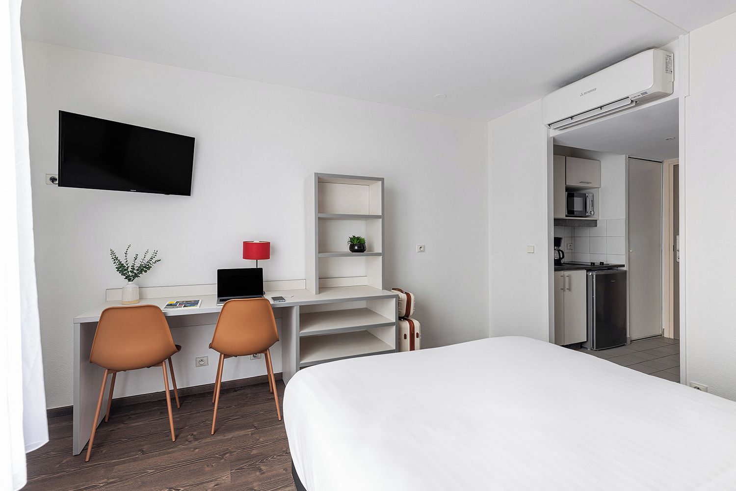 A hotel room with a bed, desk, chairs, a TV, a small shelf, and a kitchenette.