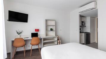 A hotel room with a bed, desk, chairs, a TV, a small shelf, and a kitchenette.