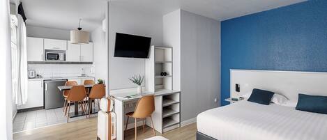 A modern hotel room with a bed, desk, and kitchenette.