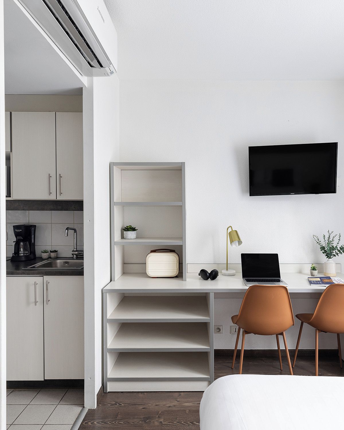 A modern hotel room with a kitchenette, a flat-screen TV mounted on the wall, a desk with a laptop, and two chairs.