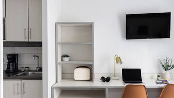 A modern hotel room with a kitchenette, a flat-screen TV mounted on the wall, a desk with a laptop, and two chairs.