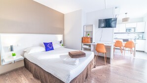 A modern hotel room with a large bed, a desk with a chair, a TV, and a kitchenette in the background. - Montempô Lyon Part Dieu (Lyon)