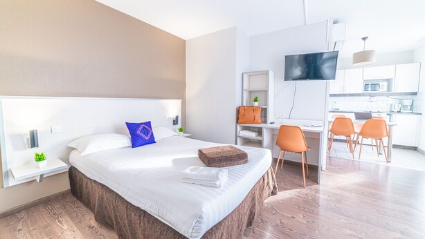 Una habitación de hotel moderna con una cama grande, un escritorio con una silla, un televisor y una kitchenette al fondo.