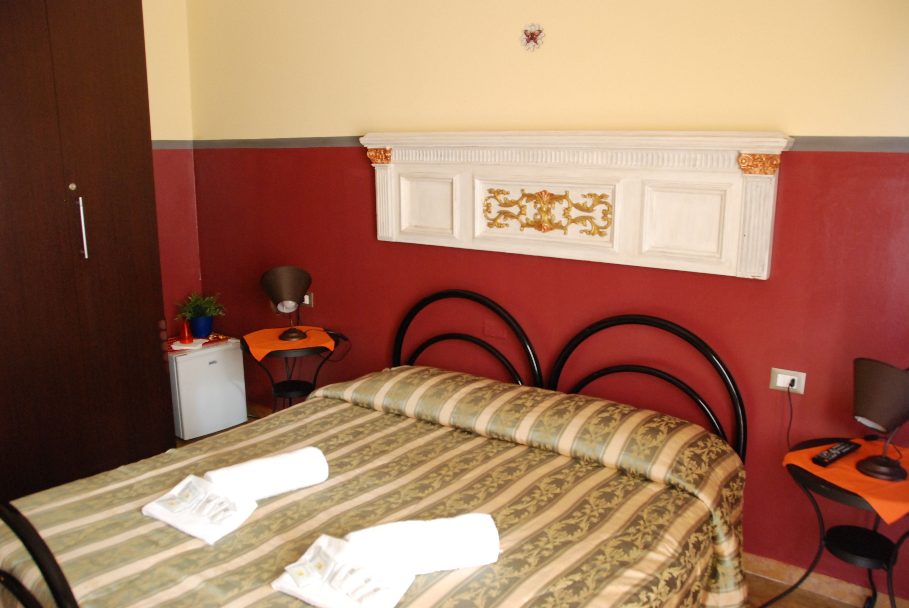 A hotel room with a bed, a desk, a chair, a small table, a potted plant, and a wall-mounted shelf.