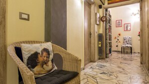 A wicker chair with a cushion and a pillow with a painting, a tiled hallway, a door, and a painting on the wall. - Soggiorno Fortezza Fiorentina (Florence)