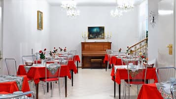 A dining hall with tables set for a meal, a piano, and a staircase leading to an upper floor.