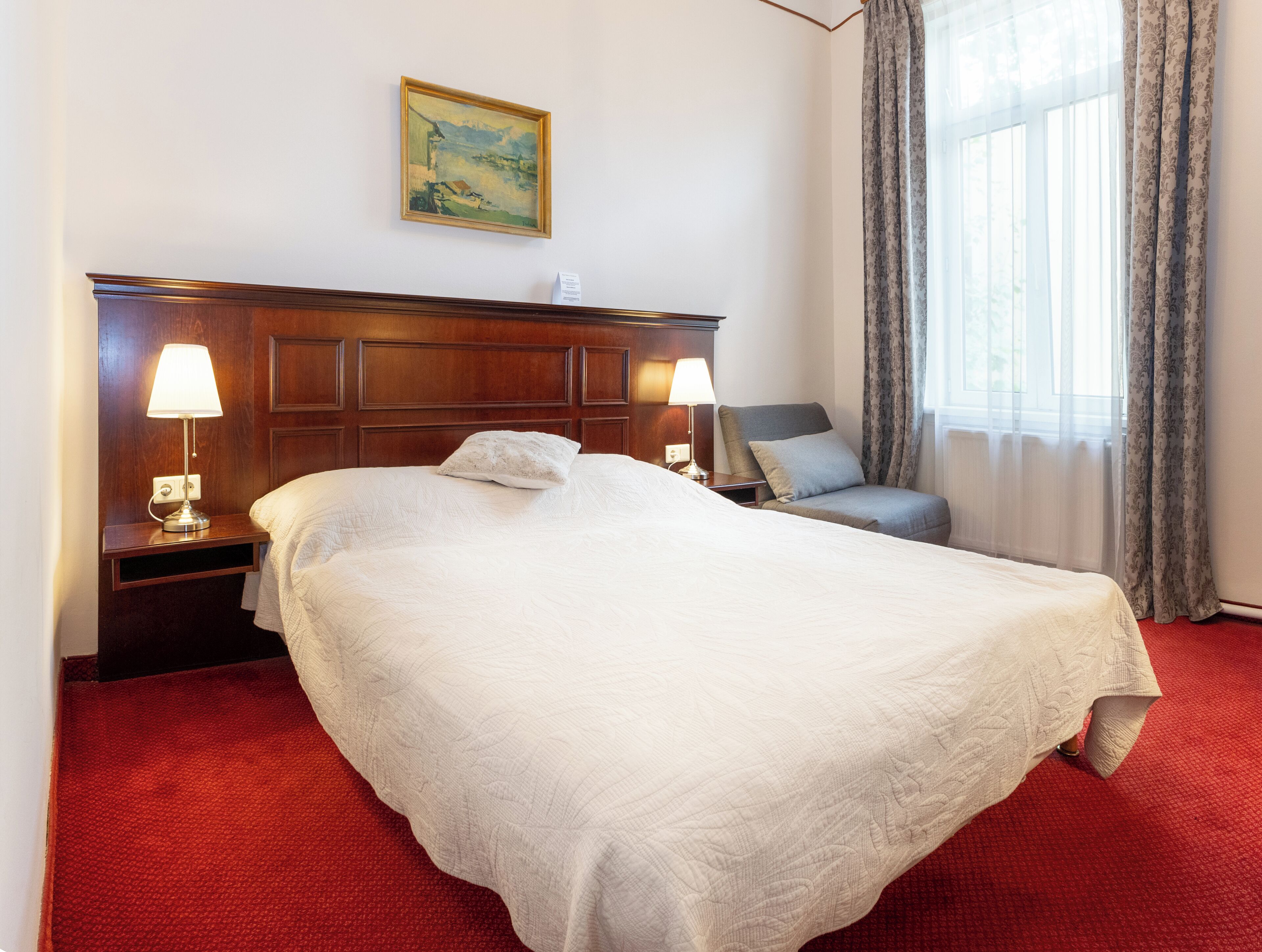Une chambre d’hôtel avec un grand lit, deux lampes de chevet, une chaise, un tableau au mur et une fenêtre avec des rideaux.