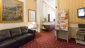 En hotellobby med en sort lædersofa, et stort blomsterbillede, en reception med en computer og en plante, samt et fjernsyn monteret på væggen.