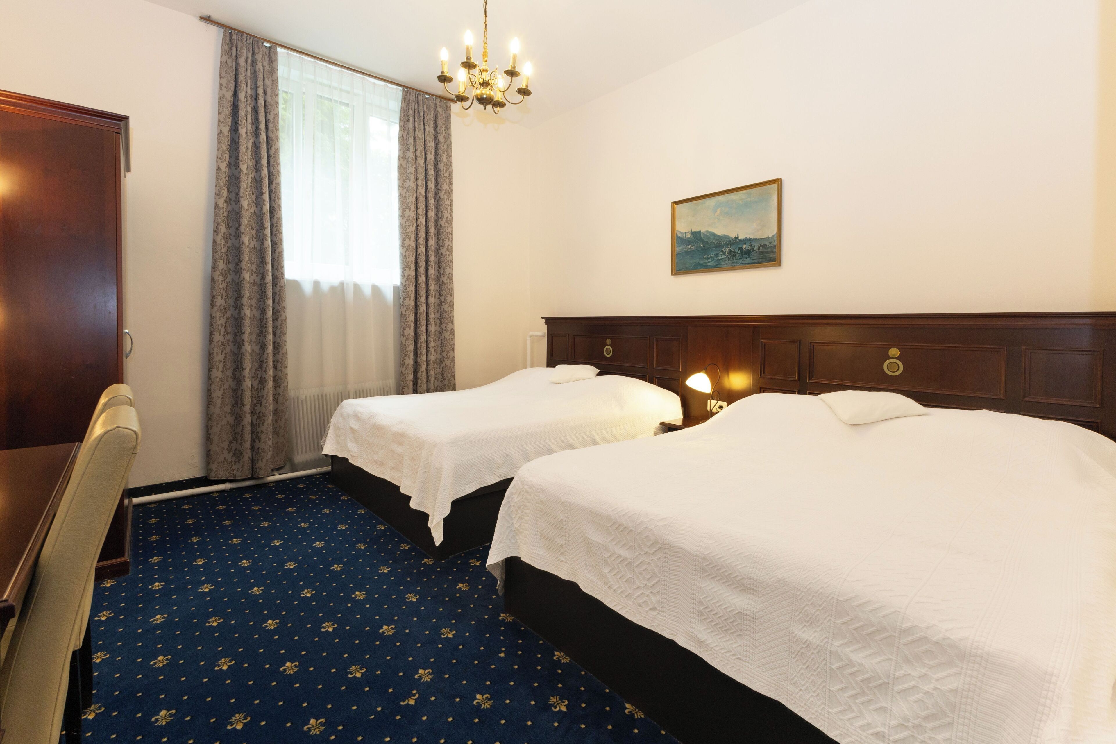 A hotel room with two beds, a chandelier, a painting on the wall, and a blue carpet with a pattern.
