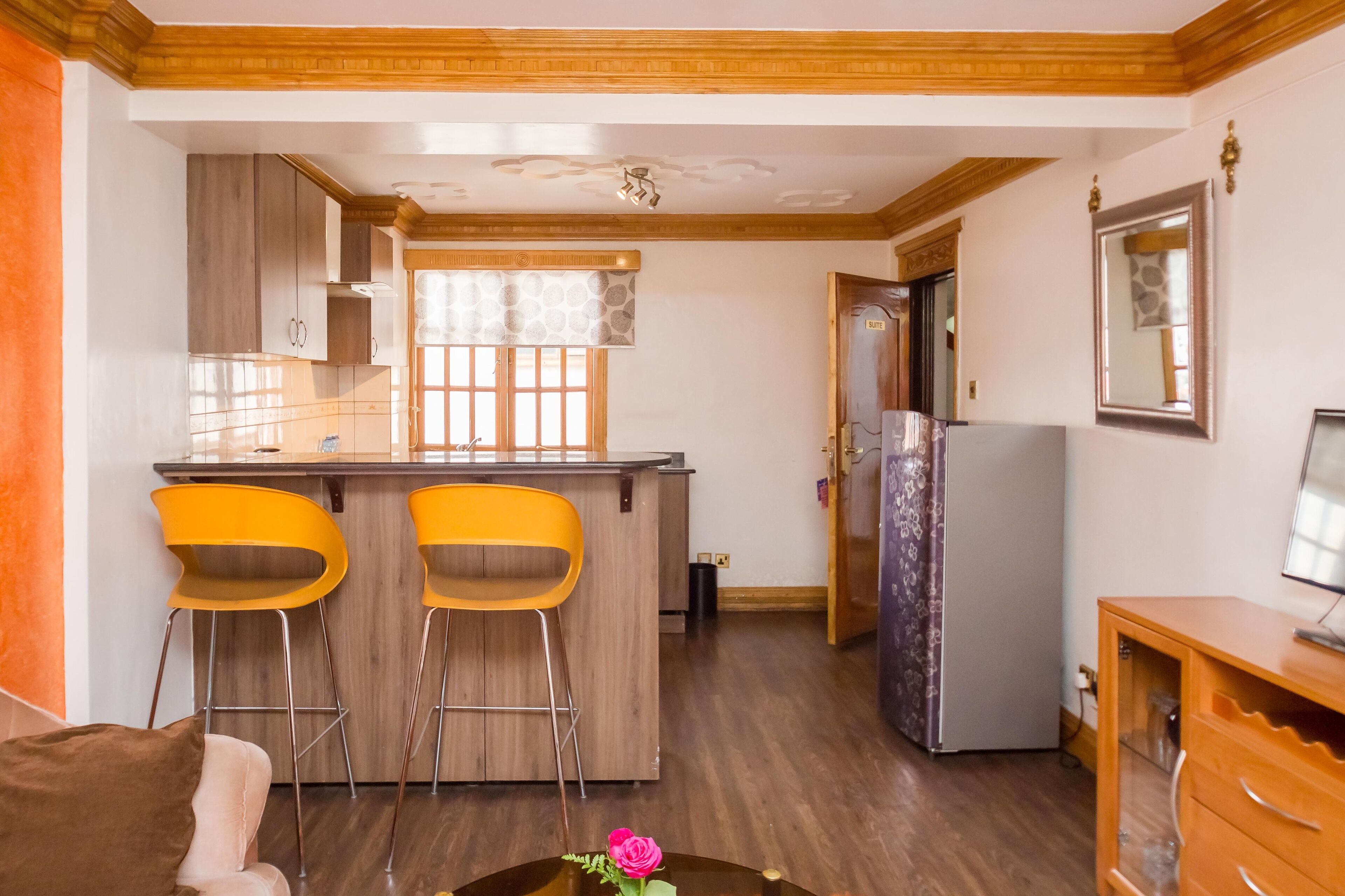 A compact kitchen with wooden cabinets, a built-in bar area with two yellow chairs, a refrigerator, and a dining area with a brown sofa and a small table with a vase.