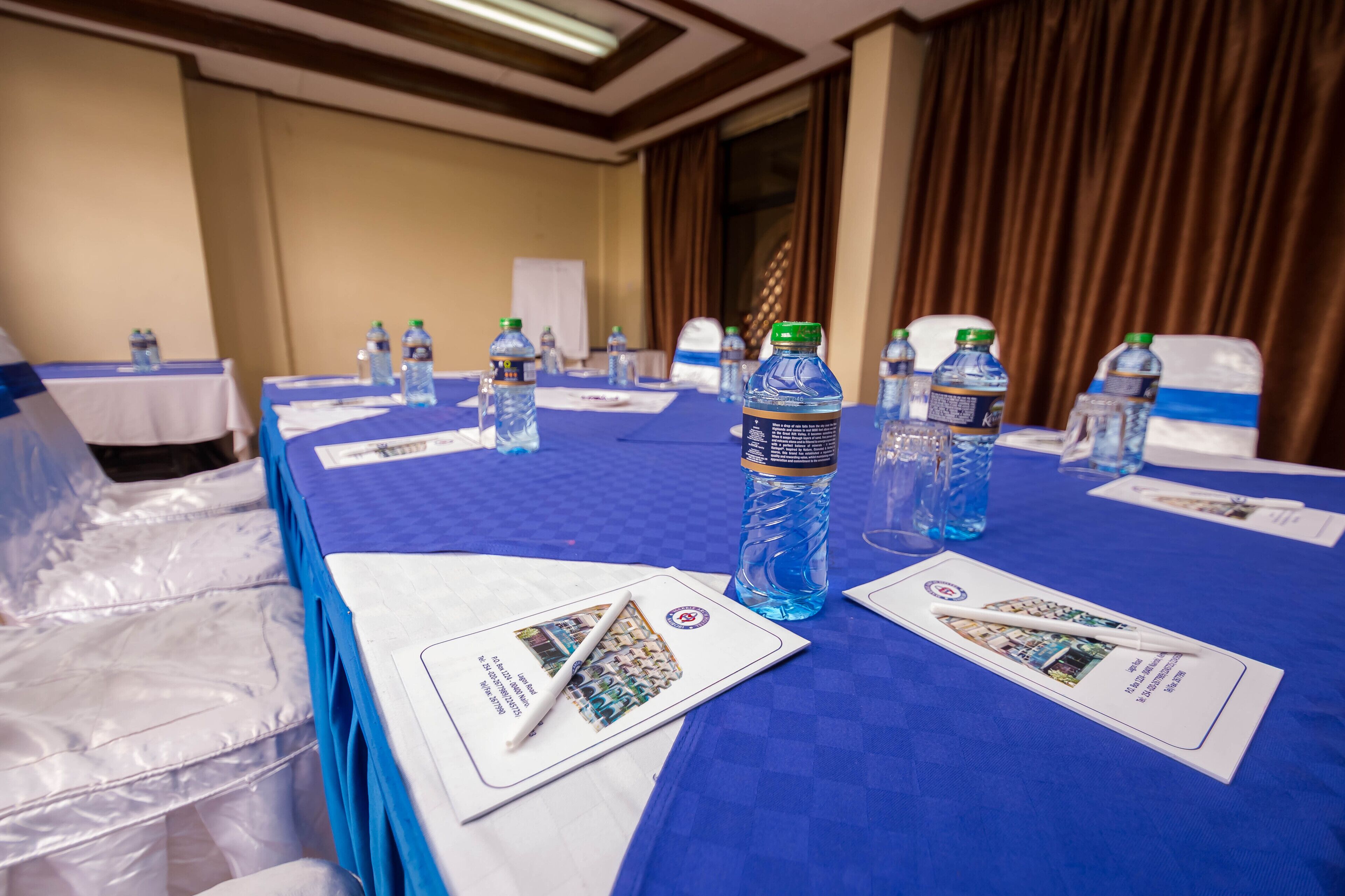 Un salón de conferencias con mesas cubiertas con manteles azules, botellas de agua y folletos informativos.