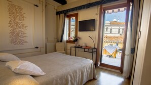 Un dormitorio con cama, escritorio y silla, televisor y balcón con vista a edificios.