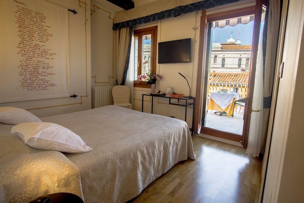 A bedroom with a bed, a desk with a chair, a TV, and a balcony with a view of buildings.