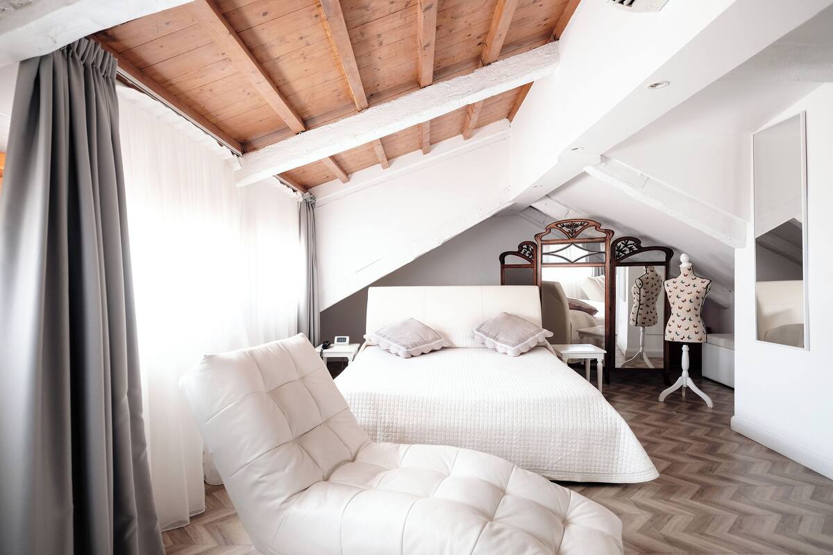 A bedroom with a loft-style ceiling, a large bed, a white armchair, and a vanity with a mirror and chair.
