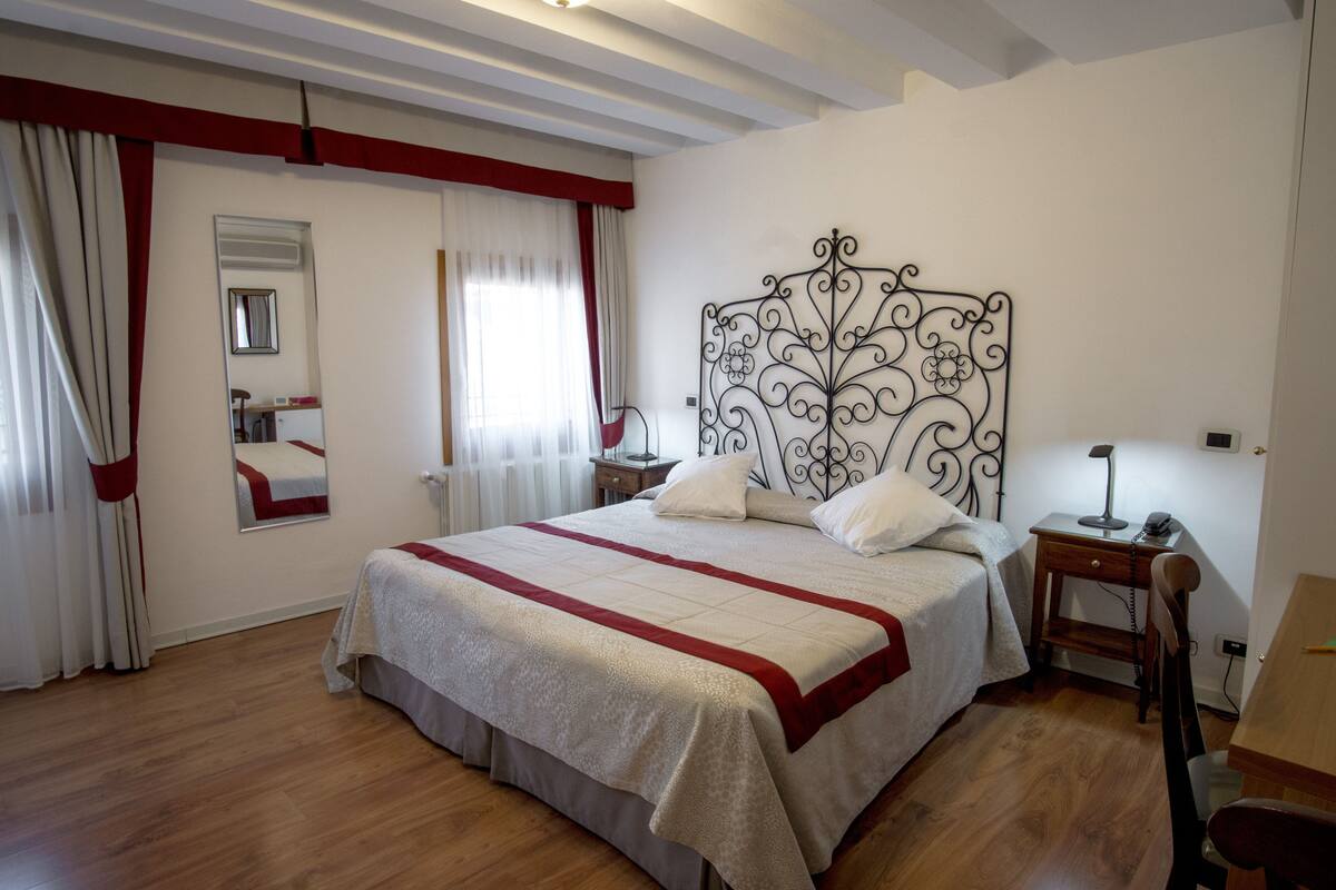 A bedroom with a large bed, a nightstand, a chair, a desk, a mirror, and a window with curtains.