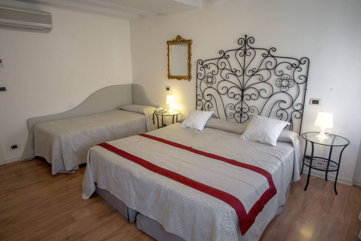 A hotel room with two beds, a decorative metal headboard, a small bedside table with a lamp, and a wall-mounted mirror.