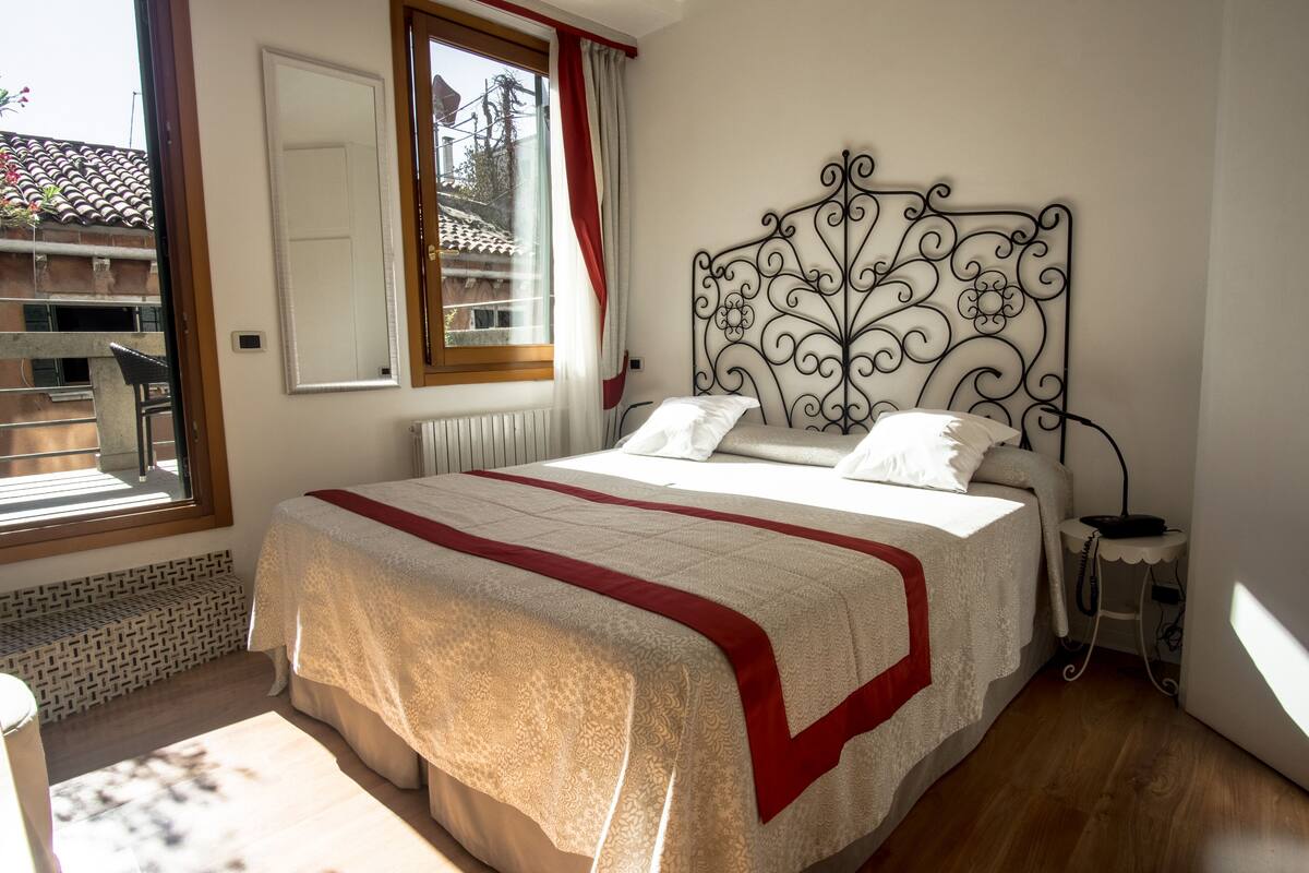 A bedroom with a bed, a window, a radiator, and a small table with a lamp.