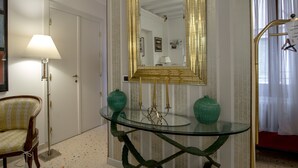 Una habitación con una mesa de vidrio, un jarrón verde, una lámpara y un espejo con marco dorado.