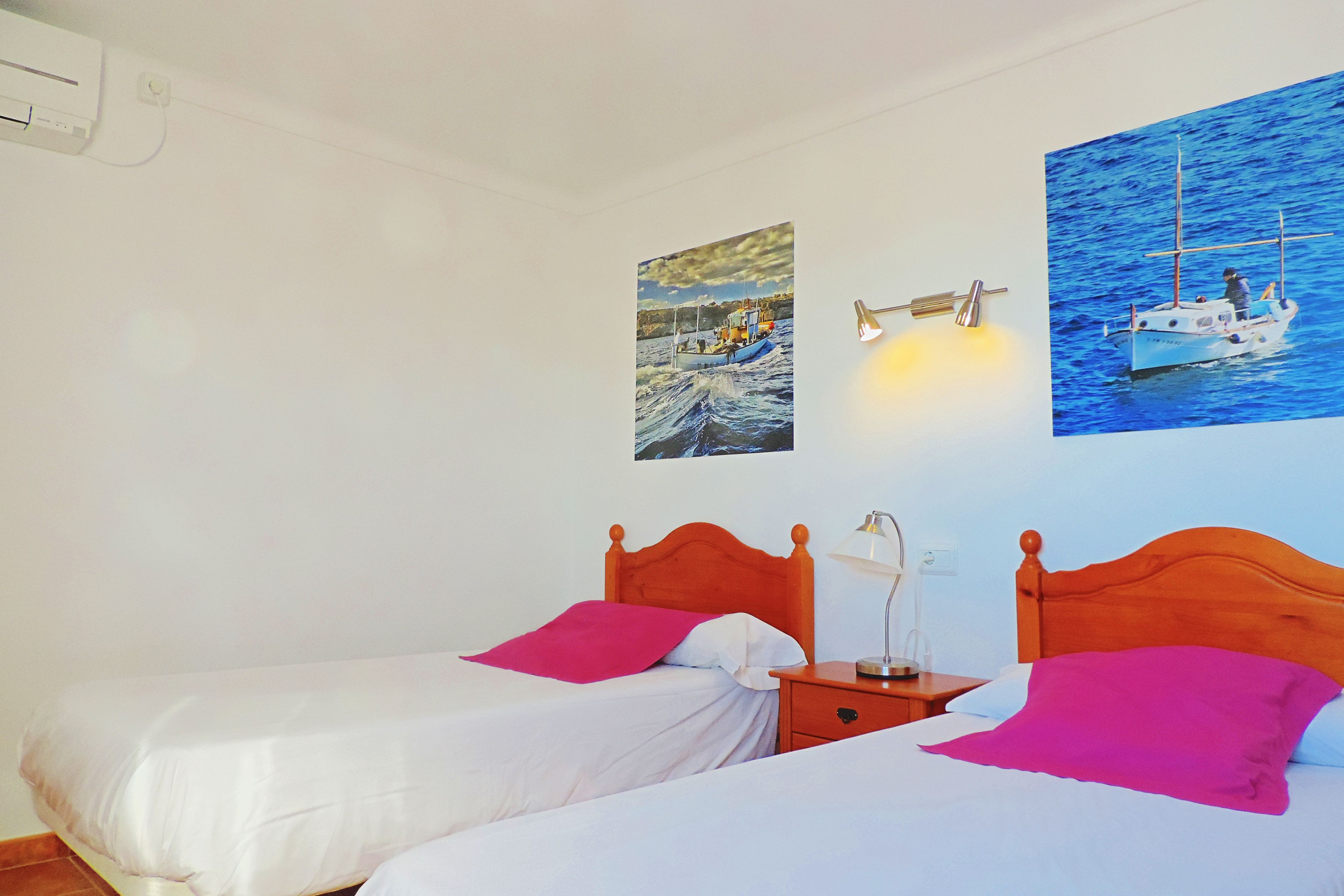 Habitación de hotel con dos camas individuales, una mesita de noche con lámpara y dos cuadros colgados en la pared con barcos.