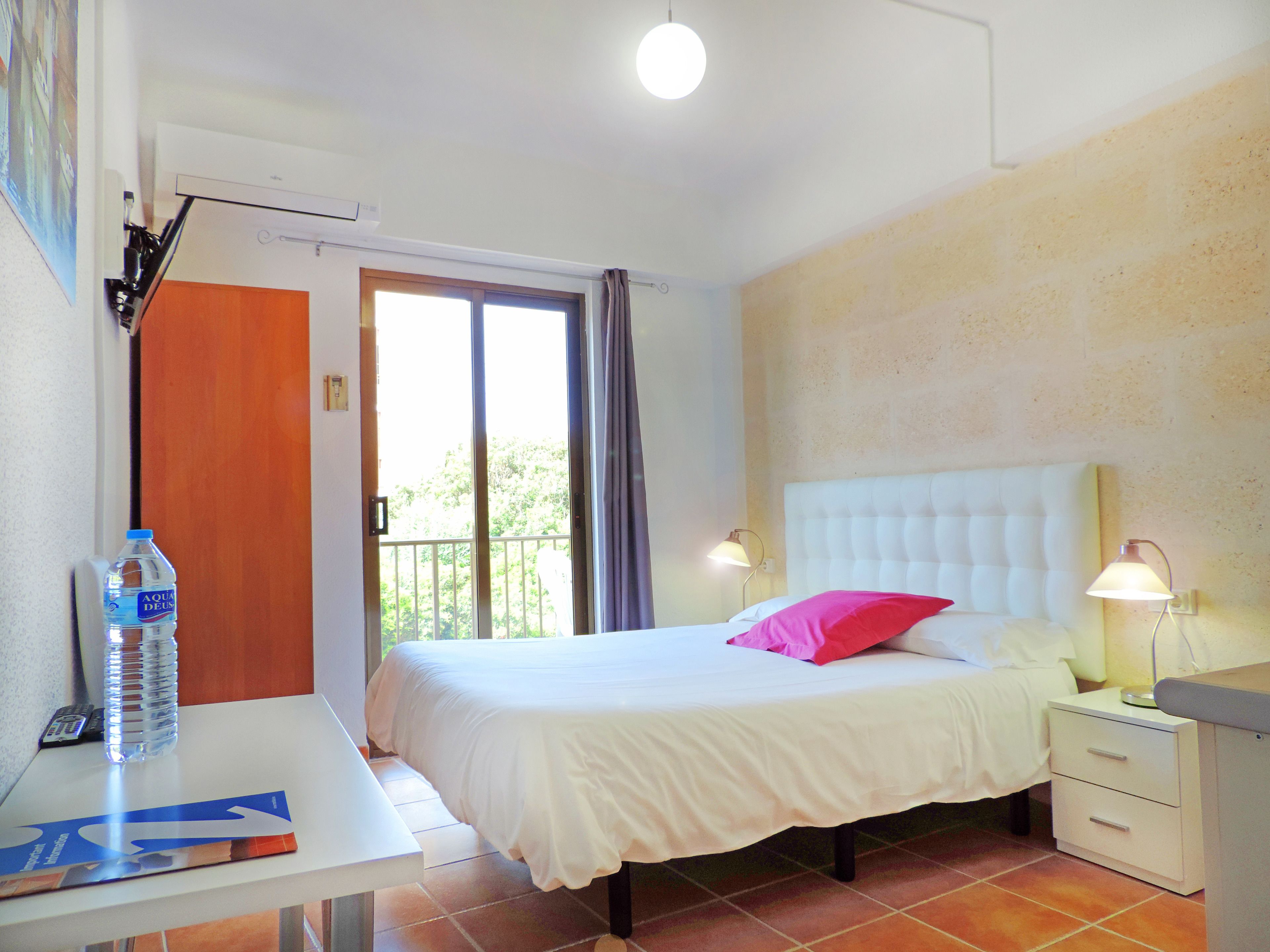 Habitación de hotel con una cama, lámparas de noche, un escritorio y una botella de agua, y vistas a través de una ventana.