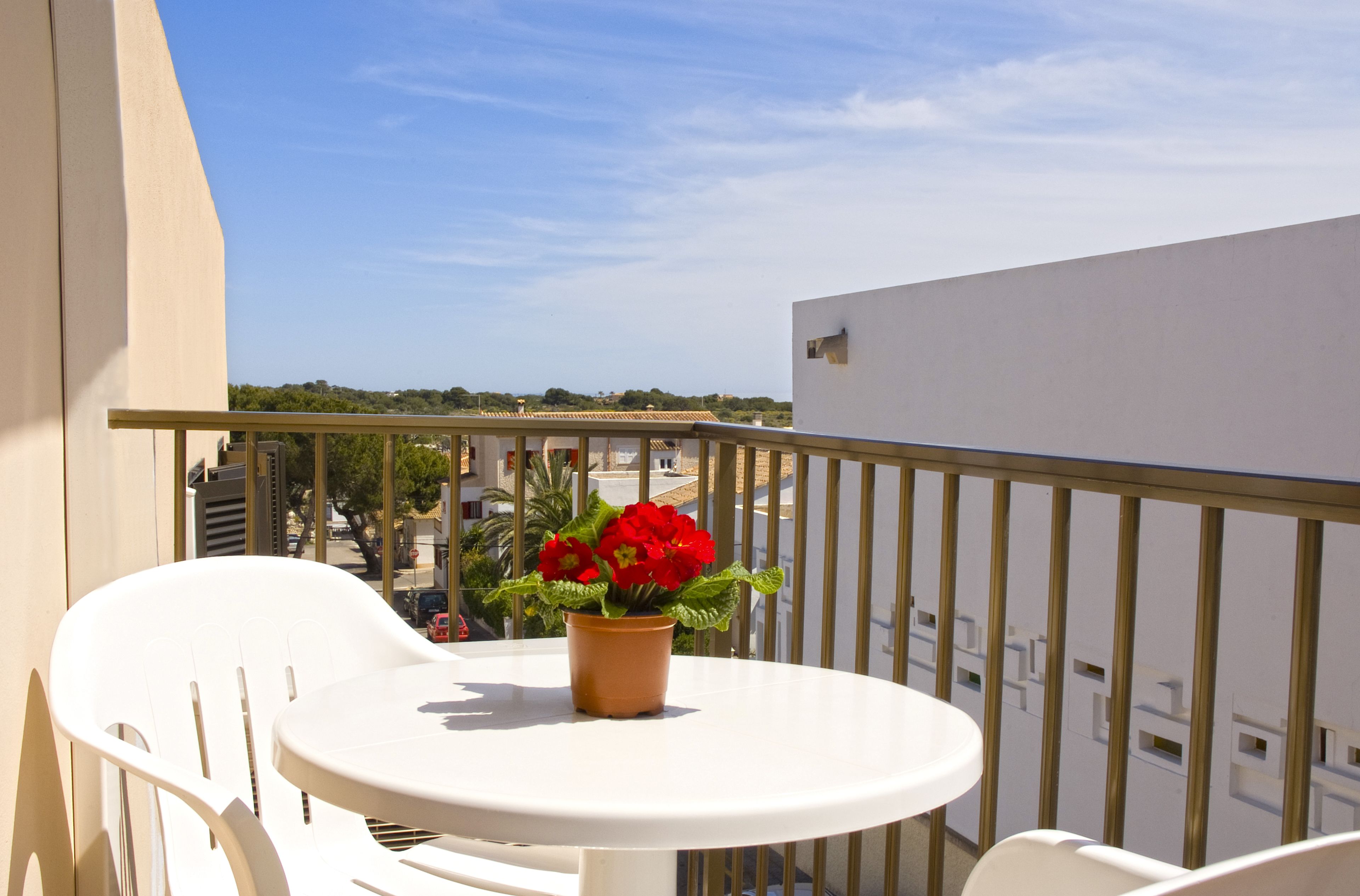 Un balcon avec une table et des chaises blanches, un pot de fleurs aux fleurs rouges, et une vue dégagée sur le paysage urbain.