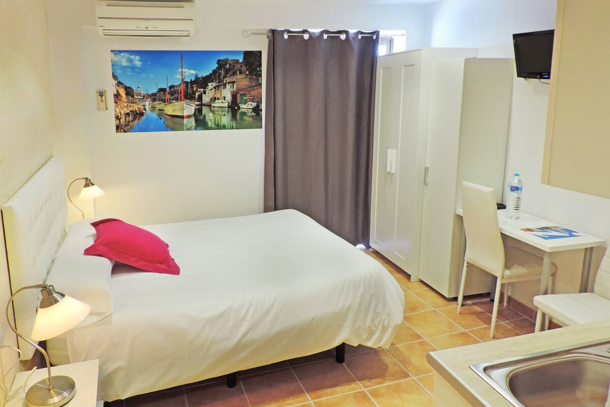 A hotel room with a bed, a desk, a chair, a sink, a mirror, a TV, and a window with curtains.