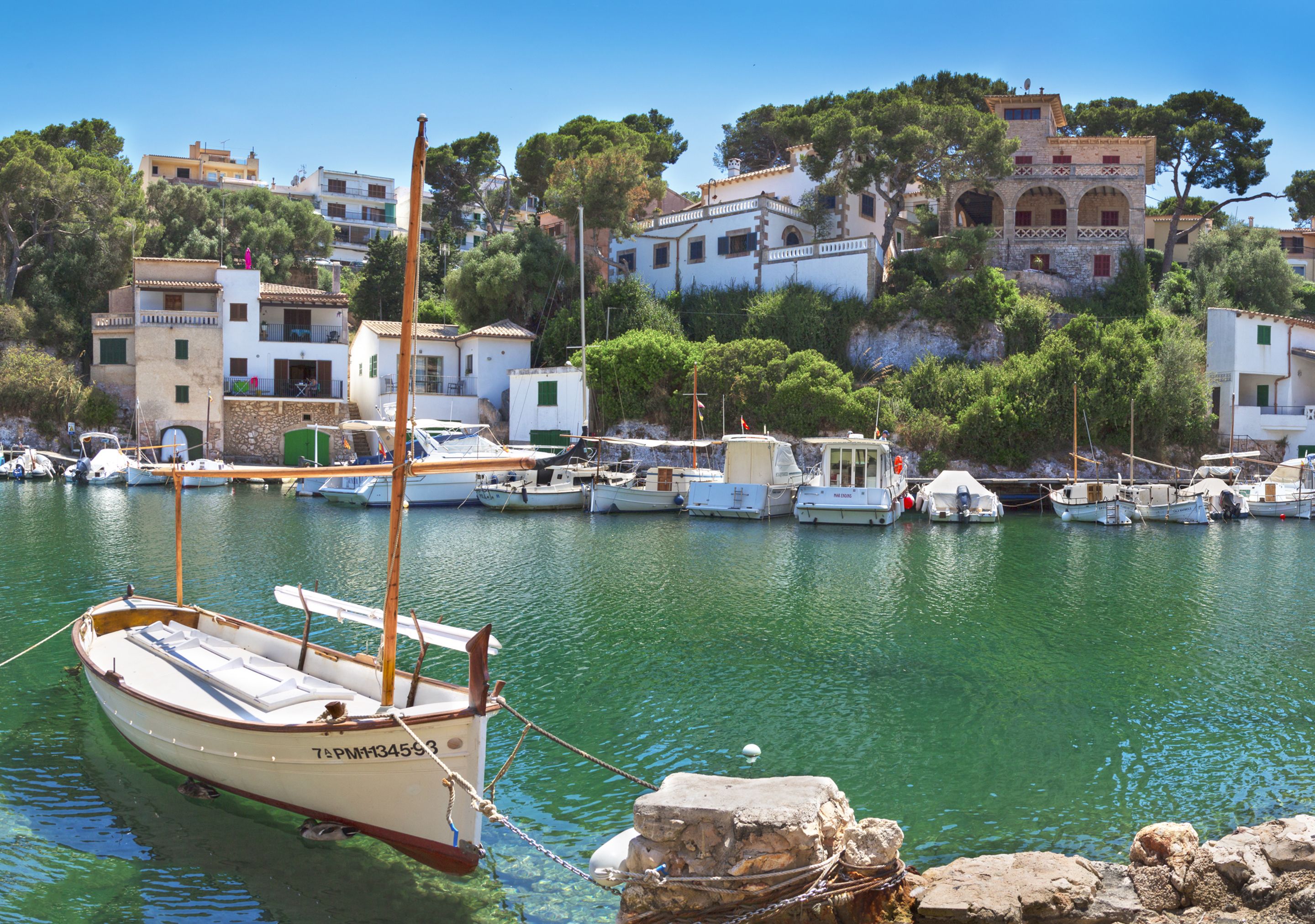 Un puerto costero con barcos amarrados, un bote pequeño en primer plano y edificios en una colina al fondo.