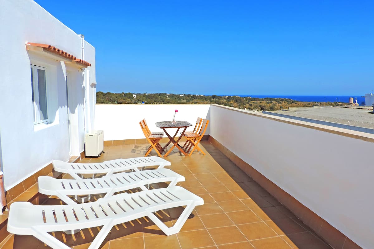 A terrace with white sun loungers, a wooden table, and chairs.