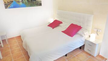 Una cama bien hecha con ropa de cama blanca y dos almohadas rojas, una mesita de noche blanca con una lámpara y suelo de baldosas.