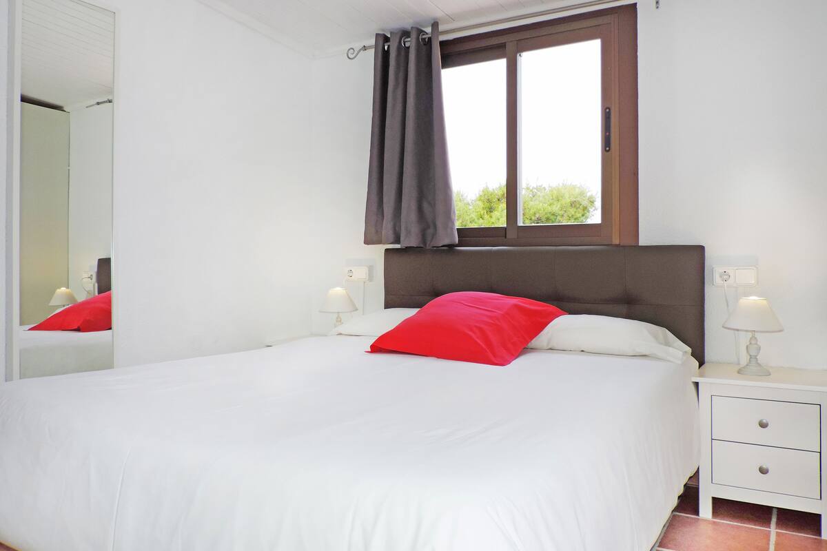 A neatly made bed with white and red bedding, a window with a curtain, a bedside lamp, and a nightstand with a drawer.