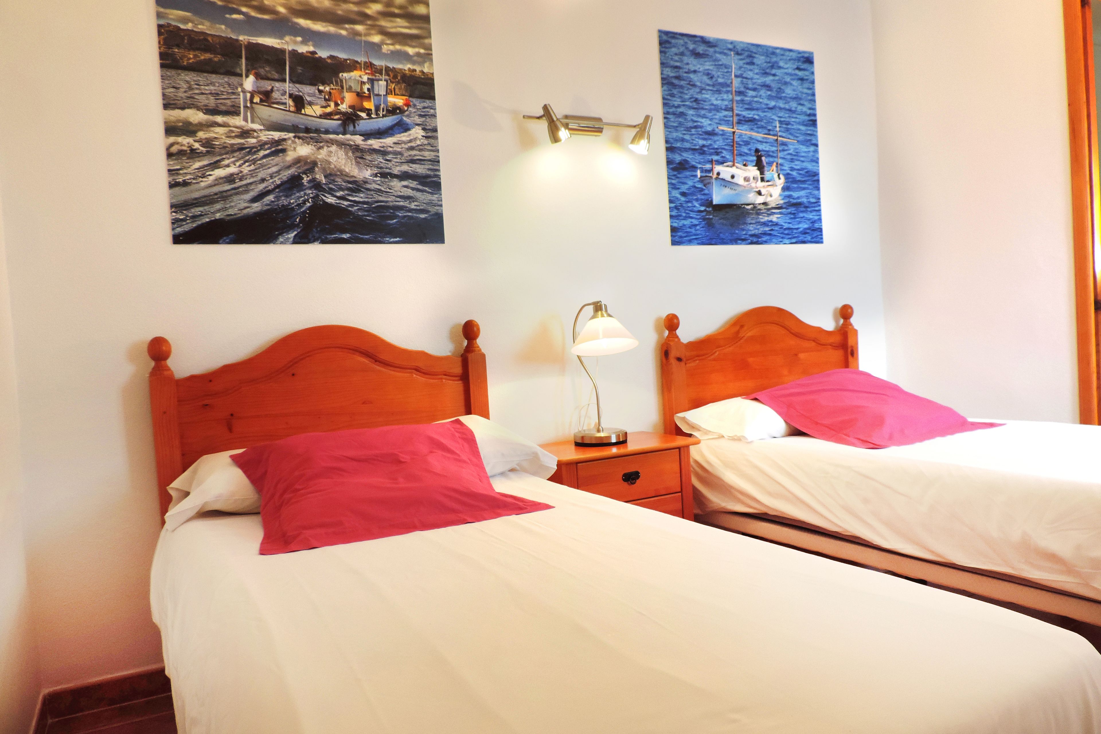 A hotel room with two beds, each with a wooden headboard, white bedding, and pink and white pillows. There are two wall-mounted lamps and two framed pictures of boats on the wall.