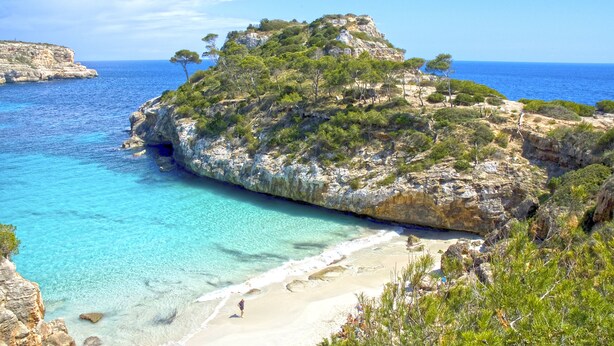 Un paysage côtier caractérisé par une colline rocheuse, une mer d’un bleu limpide et une plage de sable.