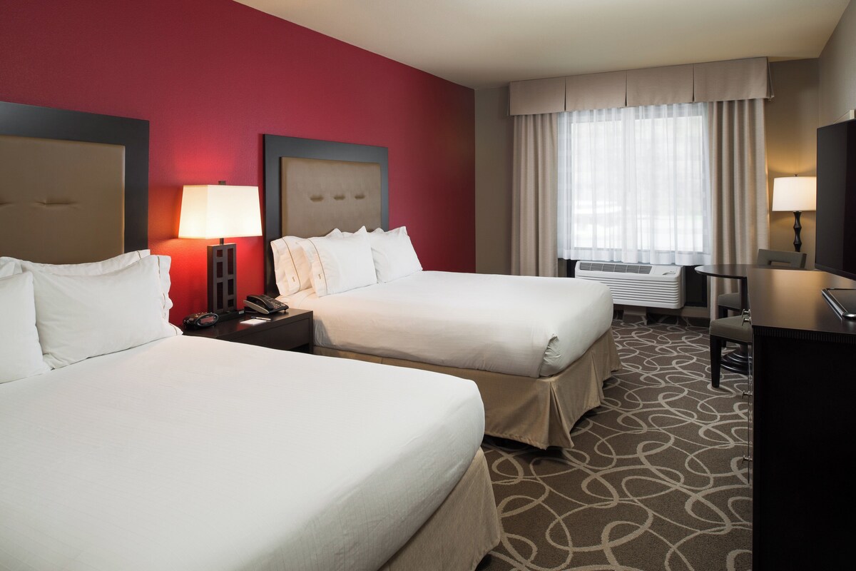 A hotel room with two beds, a desk, a TV, and a window with curtains.