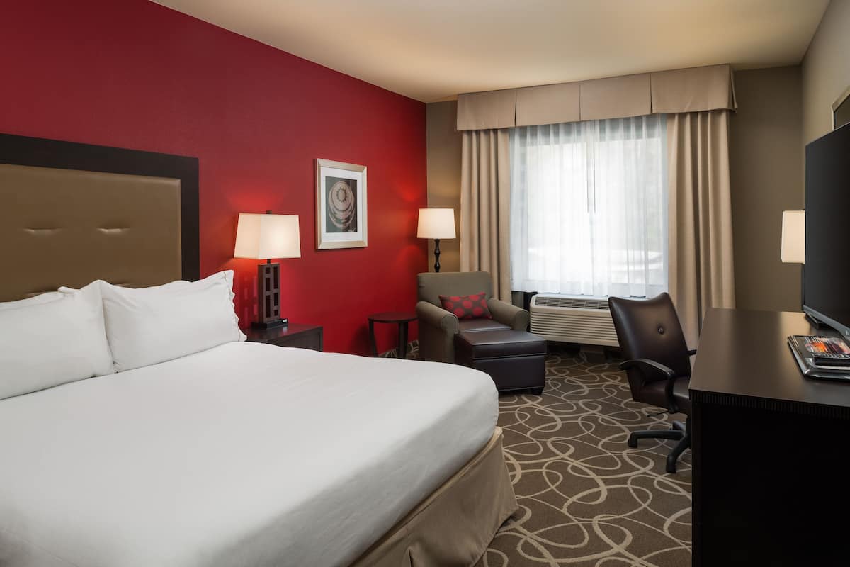 A hotel room with a large bed, two bedside tables with lamps, a desk, a chair, and a window with curtains.