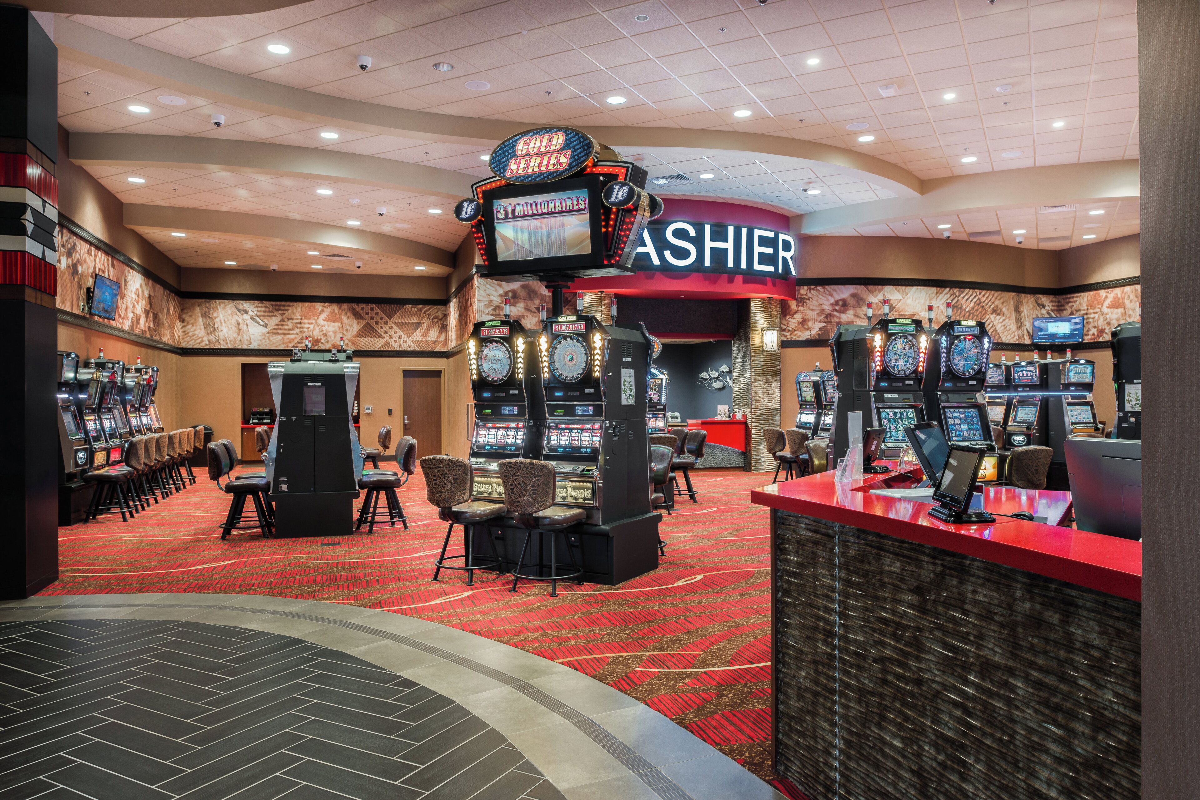 Un salón de casino con múltiples máquinas tragamonedas, un mostrador de efectivo y una zona alfombrada de color rojo.