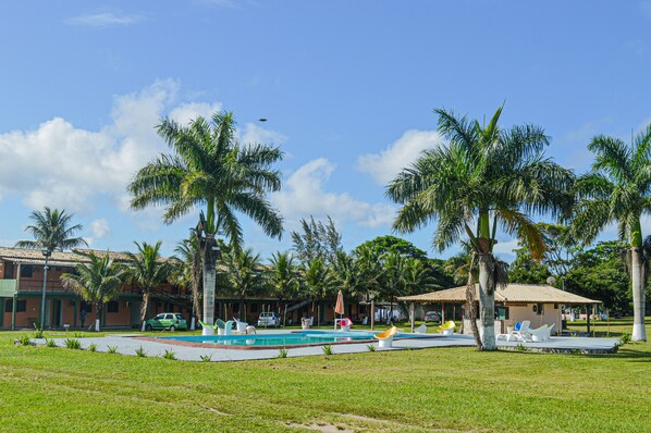 A pool area with palm trees, a small building, and a car. - Pousada Ohana (Búzios)
