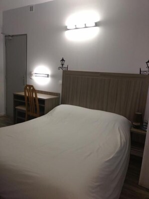 Une chambre à coucher avec un lit, un bureau et une chaise. Il y a un luminaire fixé au mur au-dessus du lit et une petite lampe de table sur le bureau.