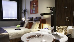 Una habitación de hotel con una cama grande, una bañera blanca con piedras, una ventana con persianas, un cuadro en la pared y una lámpara.