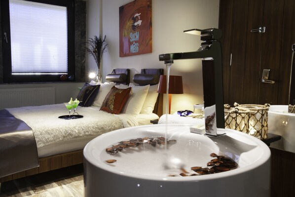Una camera d'albergo con un letto grande, una vasca da bagno bianca con pietre, una finestra con persiane, un quadro appeso al muro e una lampada.