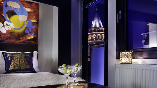 Habitación de hotel con una cama, una mesita de noche con dos vasos de martini, un cojín decorativo y vistas a una torre a través de una ventana.