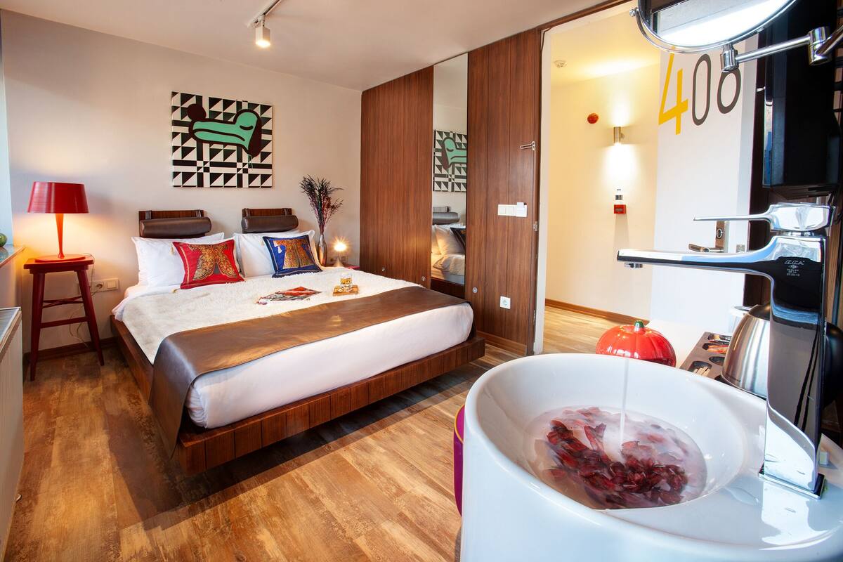 Una habitación de hotel con una cama, un espejo grande, una bañera blanca y flores rojas, y piso de madera.